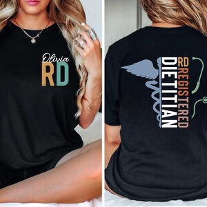 Puede incluir: Camiseta negra con el texto "Olivia RD" en blanco, turquesa y dorado. La parte trasera de la camiseta presenta las palabras "RD Registered Dietitian" con un s&iacute;mbolo de caduceo y un gr&aacute;fico de estetoscopio. La camiseta es de cuello redondo.