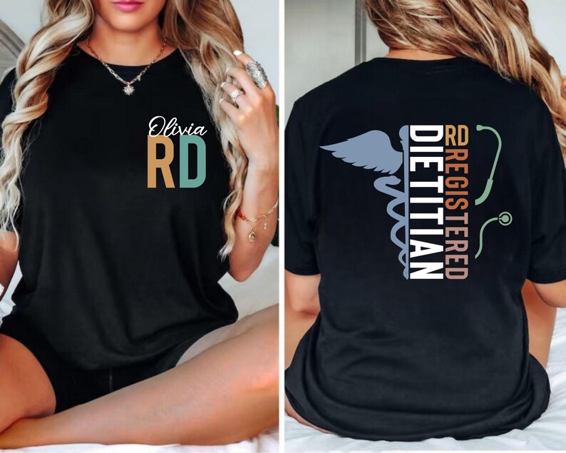Puede incluir: Camiseta negra con el texto "Olivia RD" en blanco, turquesa y dorado. La parte trasera de la camiseta presenta las palabras "RD Registered Dietitian" con un s&iacute;mbolo de caduceo y un gr&aacute;fico de estetoscopio. La camiseta es de cuello redondo.