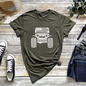 May include: A green t-shirt with a white graphic of a Jeep with an American flag on the hood. The Jeep is driving through a forest.