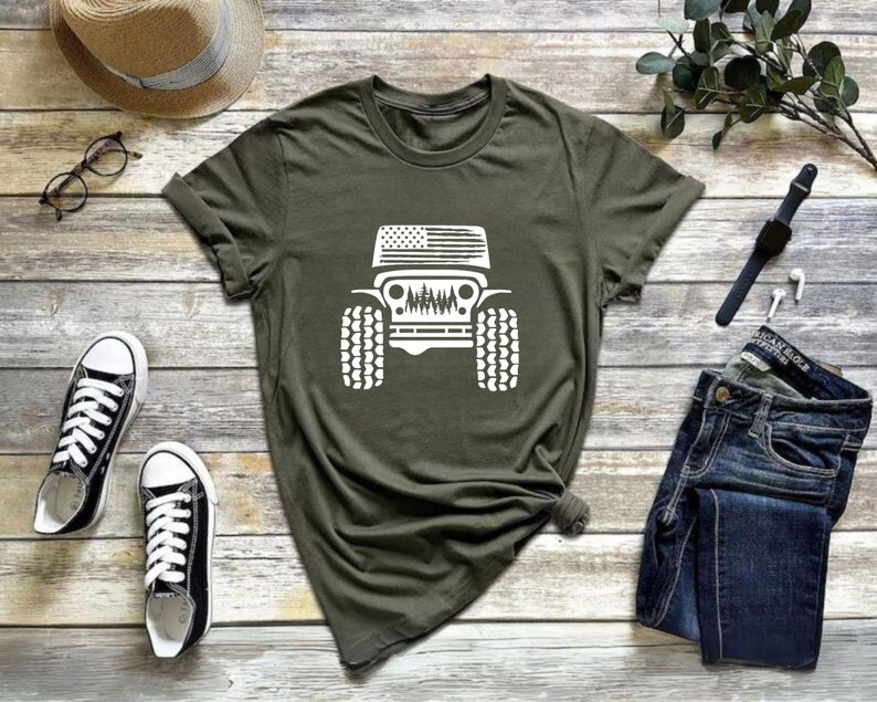 May include: A green t-shirt with a white graphic of a Jeep with an American flag on the hood. The Jeep is driving through a forest.