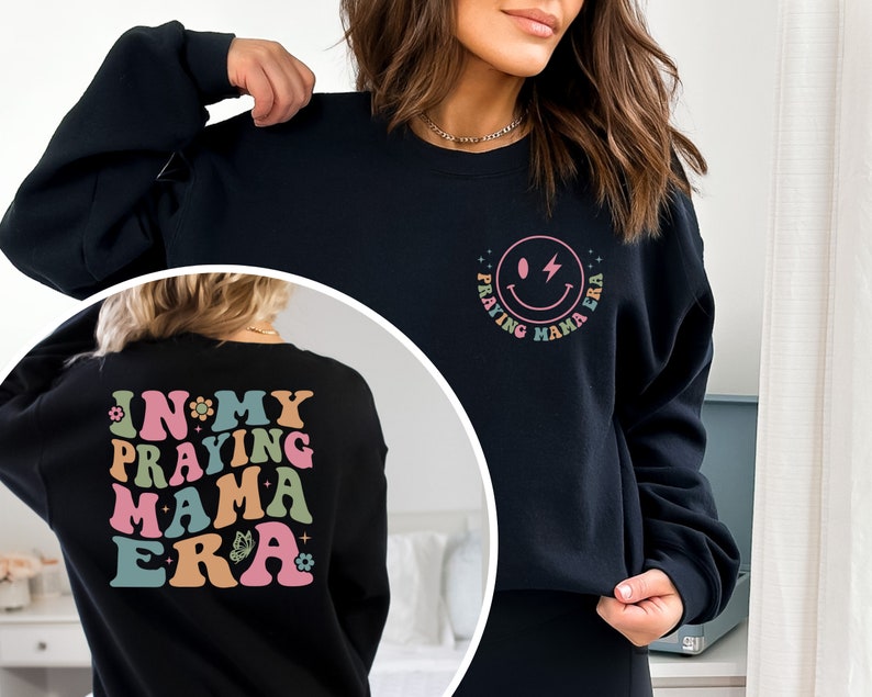 May include: Black sweatshirt with a graphic on the front that says "Praying Mama Era" with a smiley face and lightning bolt. The back of the sweatshirt has a graphic that says "In My Praying Mama Era" with flowers and a butterfly.