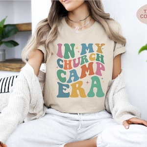 K&ouml;nnte beinhalten: Ein beiges T-Shirt mit dem Text "In My Church Camp Era" in bunten, Retro-Lettern.