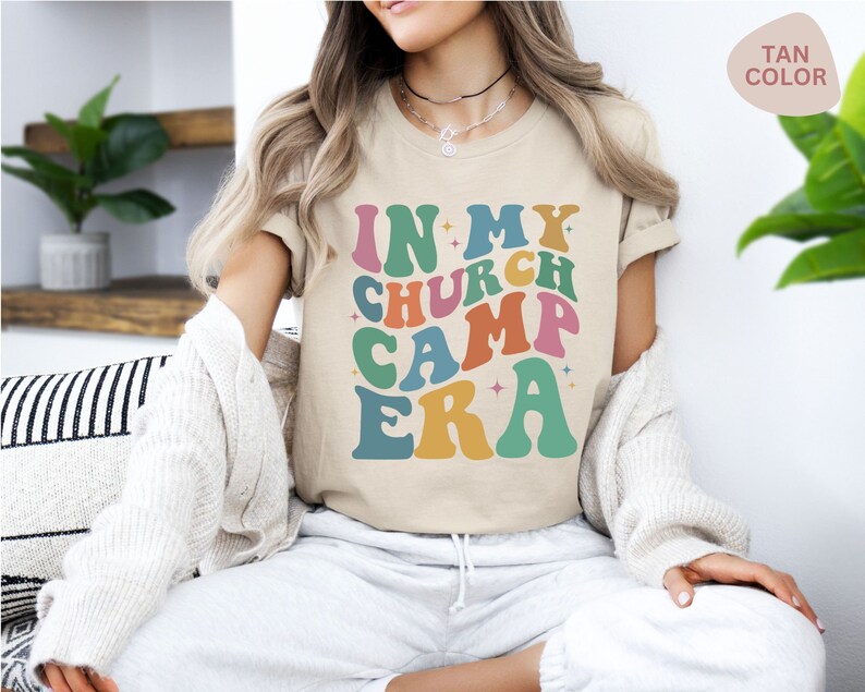 K&ouml;nnte beinhalten: Ein beiges T-Shirt mit dem Text "In My Church Camp Era" in bunten, Retro-Lettern.