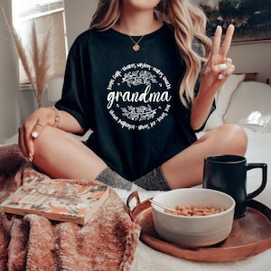 Op de afbeelding: Zwart T-shirt met het woord "grandma" in witte letters, omringd door een bloemenkrans en zinnen als "houdt geloof" en "geeft vreugde". De afbeelding bevat een dienblad met een kom eten en een zwarte mok.