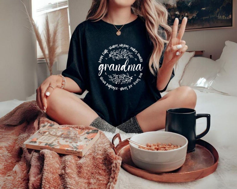 Op de afbeelding: Zwart T-shirt met het woord "grandma" in witte letters, omringd door een bloemenkrans en zinnen als "houdt geloof" en "geeft vreugde". De afbeelding bevat een dienblad met een kom eten en een zwarte mok.