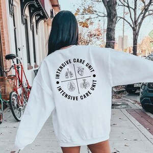 May include: A white sweatshirt with long sleeves. The back of the sweatshirt features a circular design with the words "Intensive Care Unit" and illustrations of the lungs, heart, brain, and kidneys. The sweatshirt is worn outdoors.