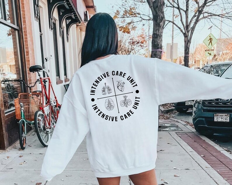 May include: A white sweatshirt with long sleeves. The back of the sweatshirt features a circular design with the words "Intensive Care Unit" and illustrations of the lungs, heart, brain, and kidneys. The sweatshirt is worn outdoors.