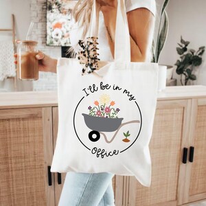May include: White tote bag with a black circle design featuring a wheelbarrow filled with colorful flowers and the text "I'll be in my office".