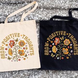 May include: Two tote bags with the text "Grow Positive Thoughts" in a retro font. The bags are made of canvas and have a floral design with butterflies. One bag is white and the other is black.