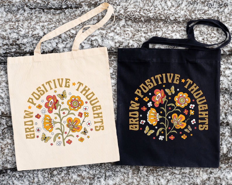 May include: Two tote bags with the text "Grow Positive Thoughts" in a retro font. The bags are made of canvas and have a floral design with butterflies. One bag is white and the other is black.