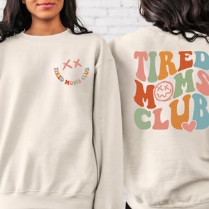 Puede incluir: Una sudadera de color crema con el texto "TIRED MOMS CLUB" en colores arco&iacute;ris en la espalda. La parte delantera de la sudadera tiene una peque&ntilde;a carita sonriente con una "X" para los ojos.