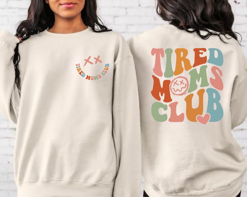 Puede incluir: Una sudadera de color crema con el texto "TIRED MOMS CLUB" en colores arco&iacute;ris en la espalda. La parte delantera de la sudadera tiene una peque&ntilde;a carita sonriente con una "X" para los ojos.
