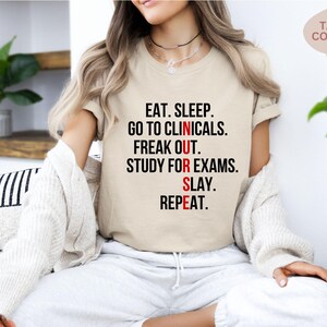 May include: Tan-colored t-shirt with the text "EAT. SLEEP. GO TO CLINICALS. FREAK OUT. STUDY FOR EXAMS. SLAY. REPEAT." in black and red. The shirt is a crew neck and has short sleeves.