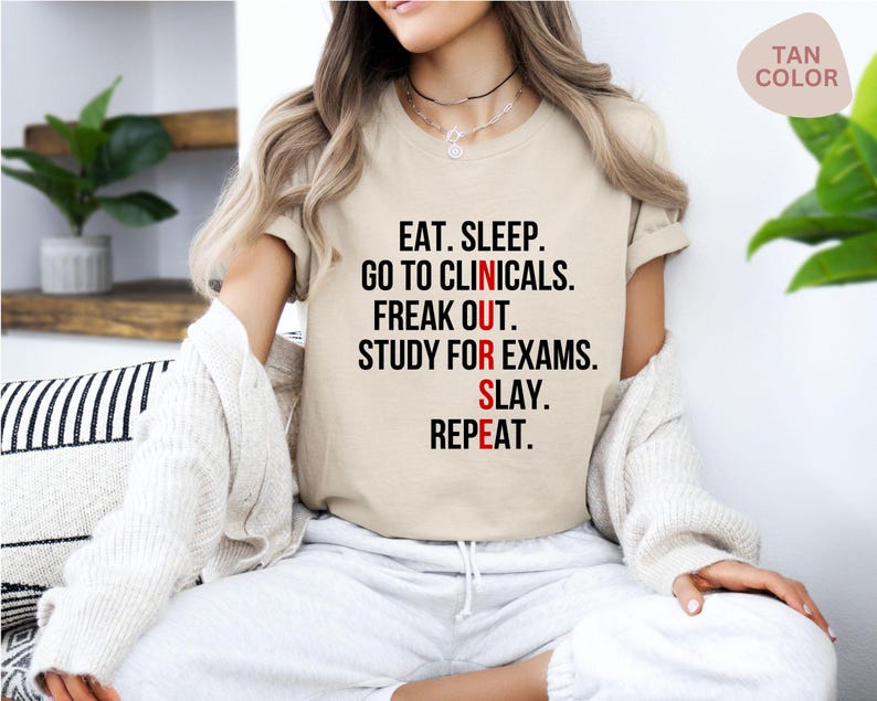 May include: Tan-colored t-shirt with the text "EAT. SLEEP. GO TO CLINICALS. FREAK OUT. STUDY FOR EXAMS. SLAY. REPEAT." in black and red. The shirt is a crew neck and has short sleeves.