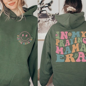 May include: A green hoodie with a smiley face graphic on the front and the text "In My Praying Mama Era" on the back. The text is in a rainbow color scheme.