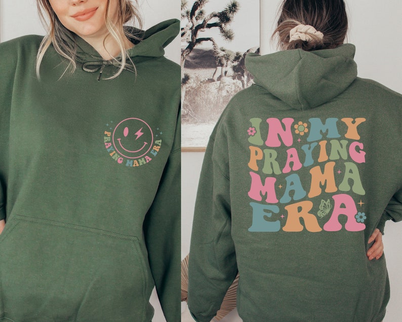 May include: A green hoodie with a smiley face graphic on the front and the text "In My Praying Mama Era" on the back. The text is in a rainbow color scheme.