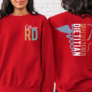 May include: Red sweatshirt with the text "Olivia RD" on the front. The back of the sweatshirt features the words "RD Registered Dietitian" with a caduceus and stethoscope graphic. The sweatshirt is a solid red color.