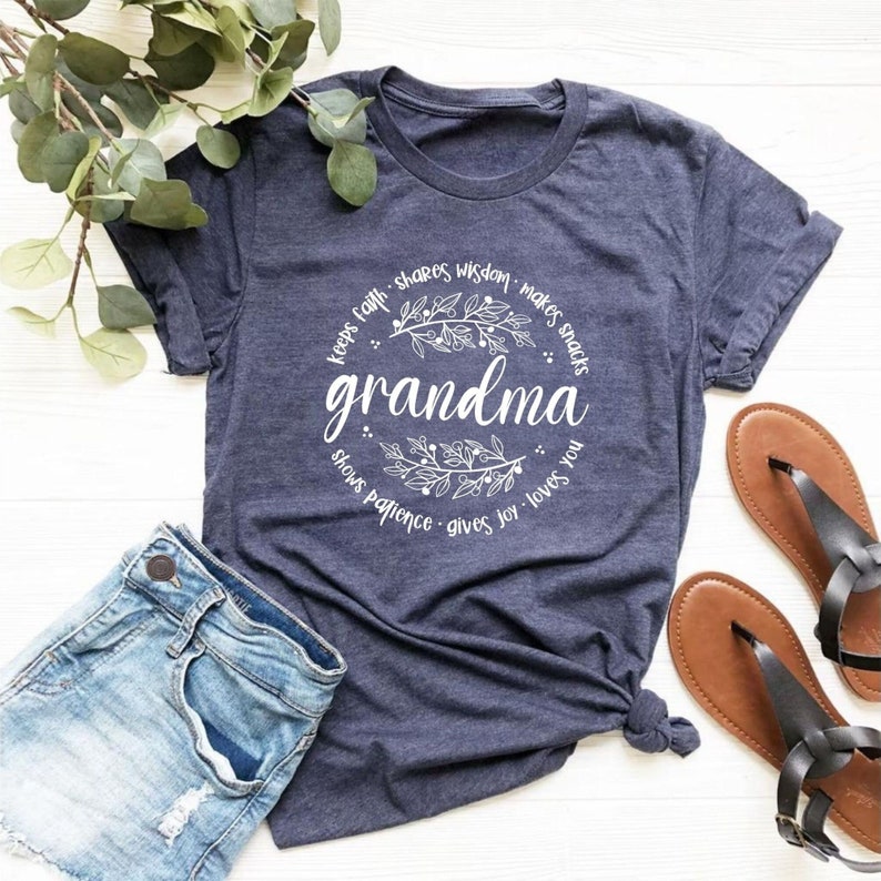 Op de afbeelding: Een marineblauw t-shirt met het woord "oma" in een wit bloemmotief. Het ontwerp bevat zinnen als "houdt geloof" en "houdt van je". Het shirt wordt gecombineerd met een spijkerbroek en bruine sandalen.