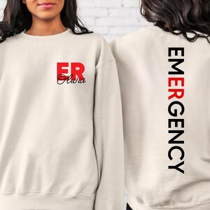 May include: A cream-colored sweatshirt with the text "ER Olivia" in red and black on the front and "EMERGENCY" in black on the back.