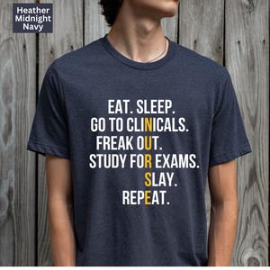 May include: Heather Midnight Navy t-shirt with the text "EAT. SLEEP. GO TO CLINICALS. FREAK OUT. STUDY FOR EXAMS. SLAY. REPEAT." in white and yellow lettering. The shirt is a crew neck style.