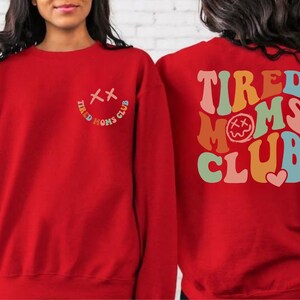 Puede incluir: Sudadera roja con el texto "Tired Moms Club" en colores arco&iacute;ris en la espalda y una peque&ntilde;a carita sonriente con una "X" por ojos en la parte delantera.