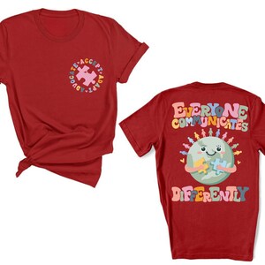 May include: Two red t-shirts with a graphic design. The front shirt has a puzzle piece with the text "Advocate, Accept, Adapt". The back shirt has a cartoon earth holding a puzzle piece with the text "Everyone Communicates Differently".