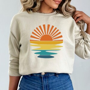 May include: Beige sweatshirt featuring a retro sunset design. The graphic includes a sun with orange rays, above horizontal bands of yellow, teal, and blue, suggesting a reflection. The sweatshirt has long sleeves and a crew neck.