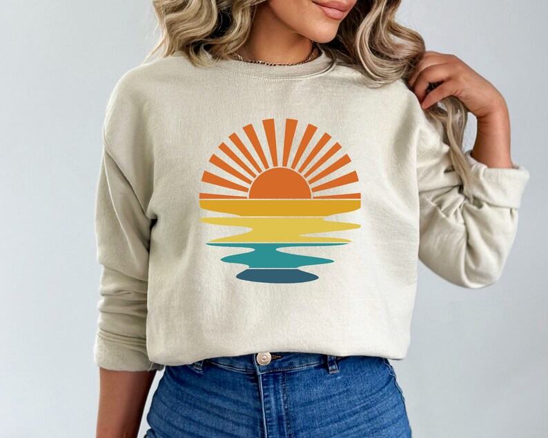 May include: Beige sweatshirt featuring a retro sunset design. The graphic includes a sun with orange rays, above horizontal bands of yellow, teal, and blue, suggesting a reflection. The sweatshirt has long sleeves and a crew neck.