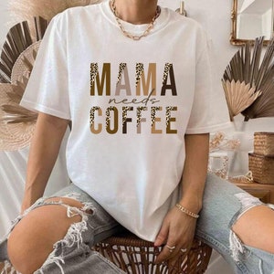 Op de afbeelding: Wit T-shirt met de tekst "MAMA needs COFFEE" in bruine en zwarte letters met een luipaardprint.