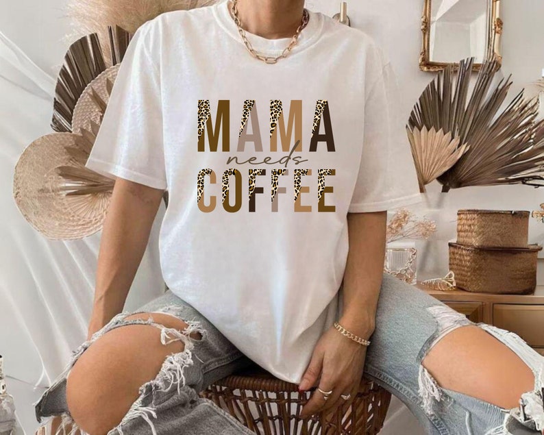 Op de afbeelding: Wit T-shirt met de tekst "MAMA needs COFFEE" in bruine en zwarte letters met een luipaardprint.