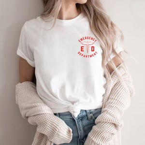 Puede incluir: Camiseta blanca con el logo rojo "Emergency Department" con un s&iacute;mbolo de caduceo. La camiseta tiene mangas cortas y cuello redondo. La modelo lleva vaqueros azules y un su&eacute;ter color crema.