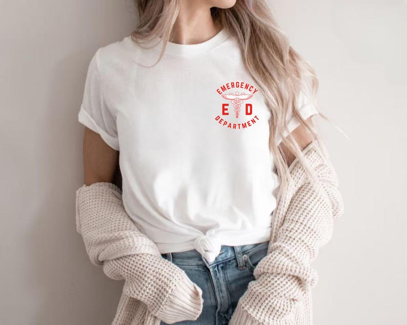 Puede incluir: Camiseta blanca con el logo rojo "Emergency Department" con un s&iacute;mbolo de caduceo. La camiseta tiene mangas cortas y cuello redondo. La modelo lleva vaqueros azules y un su&eacute;ter color crema.