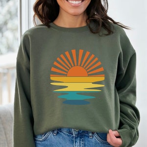 May include: Olive green sweatshirt featuring a retro sunset design. The graphic includes a vibrant orange sun with radiating lines, set above yellow, orange, and blue water reflections. A casual, comfortable top.