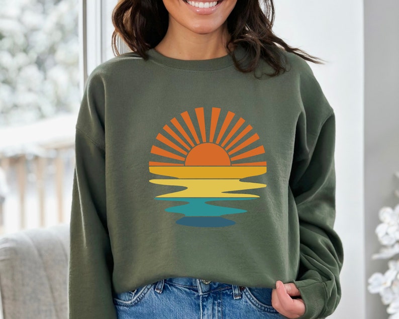 May include: Olive green sweatshirt featuring a retro sunset design. The graphic includes a vibrant orange sun with radiating lines, set above yellow, orange, and blue water reflections. A casual, comfortable top.