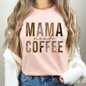 Op de afbeelding: Een roze T-shirt met de tekst "MAMA needs COFFEE" in bruine, beige en zwarte letters met een luipaardprint.