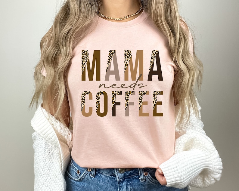 Op de afbeelding: Een roze T-shirt met de tekst "MAMA needs COFFEE" in bruine, beige en zwarte letters met een luipaardprint.