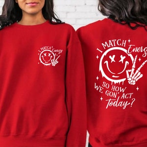 Puede incluir: Sudadera roja con un estampado gr&aacute;fico blanco de una cara sonriente con los ojos cruzados y una mano de esqueleto haciendo el signo de la paz. El texto dice "I Match Energy So How We Gon' Act Today?"