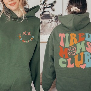 Puede incluir: Sudadera con capucha verde con un gr&aacute;fico colorido de "Tired Moms Club" en la espalda y una carita sonriente con el texto "Tired Moms Club" en la parte delantera.