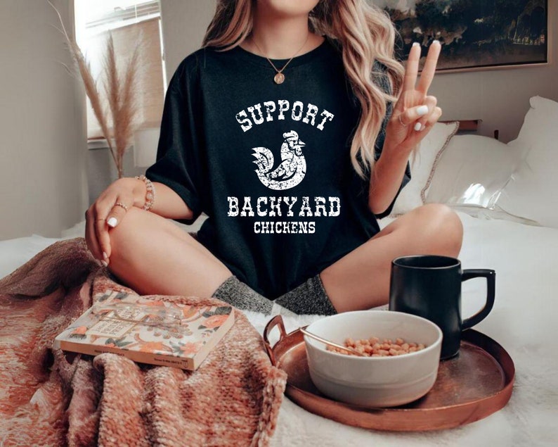 May include: A woman wearing a black t-shirt with white text that reads "Support Backyard Chickens" and a white rooster graphic. She is sitting on a bed with a blanket and a book.