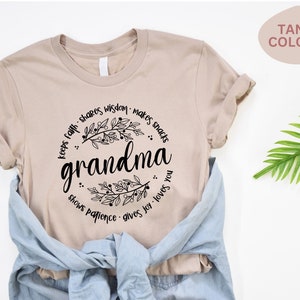 Op de afbeelding: T-shirt in een tan kleur met het woord "grandma" in zwart script, omringd door een bloemenontwerp en zinnen als "deelt wijsheid" en "houdt van je". Een blauw spijkeroverhemd is over het shirt gedrapeerd.