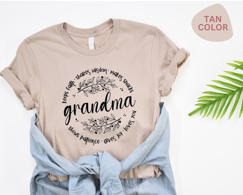 Op de afbeelding: T-shirt in een tan kleur met het woord "grandma" in zwart script, omringd door een bloemenontwerp en zinnen als "deelt wijsheid" en "houdt van je". Een blauw spijkeroverhemd is over het shirt gedrapeerd.