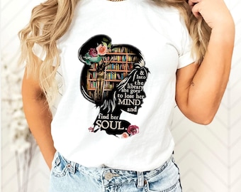 Puede incluir: Una camiseta blanca de manga corta con un gráfico de silueta negra de una cabeza llena con una escena de estantería de biblioteca, detalles florales y el texto: & Into the library she goes to lose her MIND and find her SOUL.