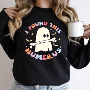 May include: Black sweatshirt with a colorful graphic that reads "I FOUND THIS HUMERUS." The design features a cartoon ghost holding a bone, with stars and bats. The text is in a retro font with pink, orange, yellow, and blue colors.