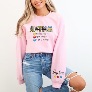 May include: Pink sweatshirt with the text "Autism Mama" in colorful puzzle piece letters. The text continues with "totally blessed, often stressed, a bit of a mess". The sweatshirt has the name "Sophia" on the sleeve with puzzle pieces.
