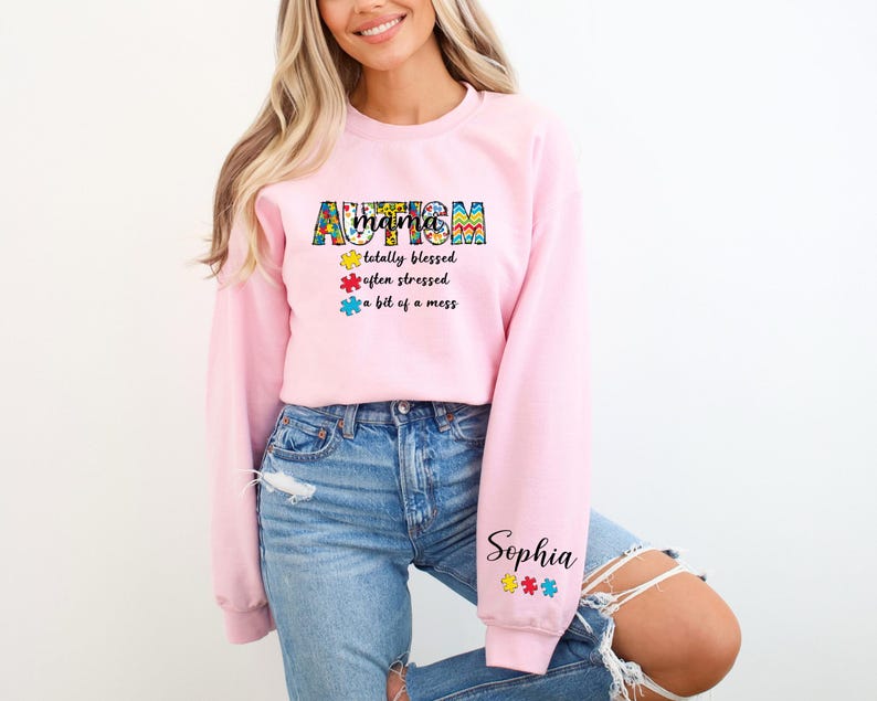 May include: Pink sweatshirt with the text "Autism Mama" in colorful puzzle piece letters. The text continues with "totally blessed, often stressed, a bit of a mess". The sweatshirt has the name "Sophia" on the sleeve with puzzle pieces.