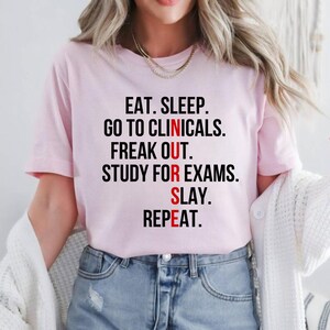 May include: A light pink t-shirt with the text "EAT. SLEEP. GO TO CLINICALS. FREAK OUT. STUDY FOR EXAMS. SLAY. REPEAT." in black and red lettering. The shirt is a crew neck style.
