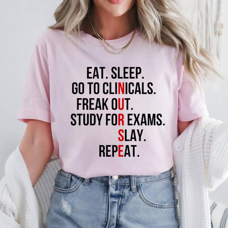 May include: A light pink t-shirt with the text "EAT. SLEEP. GO TO CLINICALS. FREAK OUT. STUDY FOR EXAMS. SLAY. REPEAT." in black and red lettering. The shirt is a crew neck style.
