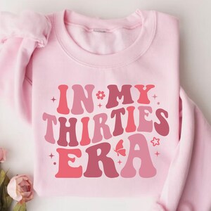 May include: Pink sweatshirt with the text "IN MY THIRTIES ERA" in a retro font with pink, red, and white colors.