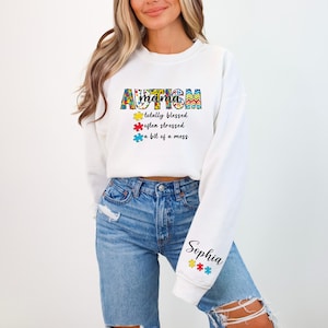 May include: White cropped sweatshirt with the text "mama autism" in colorful puzzle piece letters. The text below reads "totally blessed, often stressed, a bit of a mess". The sleeve has the name "Sophia" in black text with colorful puzzle pieces.
