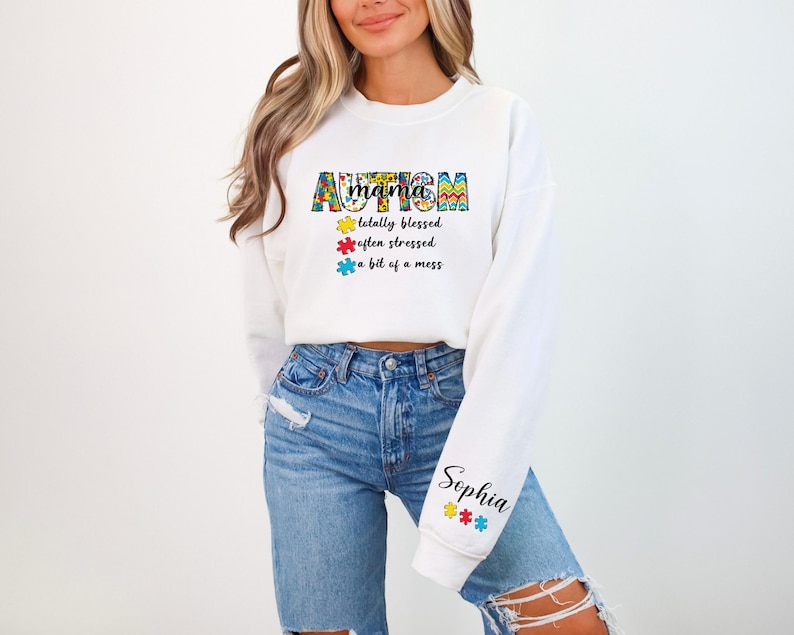 May include: White cropped sweatshirt with the text "mama autism" in colorful puzzle piece letters. The text below reads "totally blessed, often stressed, a bit of a mess". The sleeve has the name "Sophia" in black text with colorful puzzle pieces.